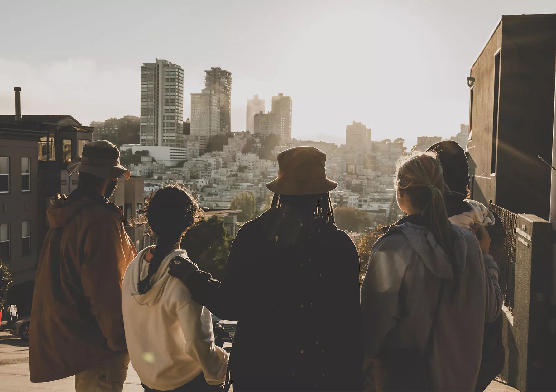 Cinco amigos parados juntos y con chaquetas y sombreros de Timberland, que se muestran desde atrás. Con el sol iluminando sus siluetas mientras miran a una ciudad con edificios blancos.
