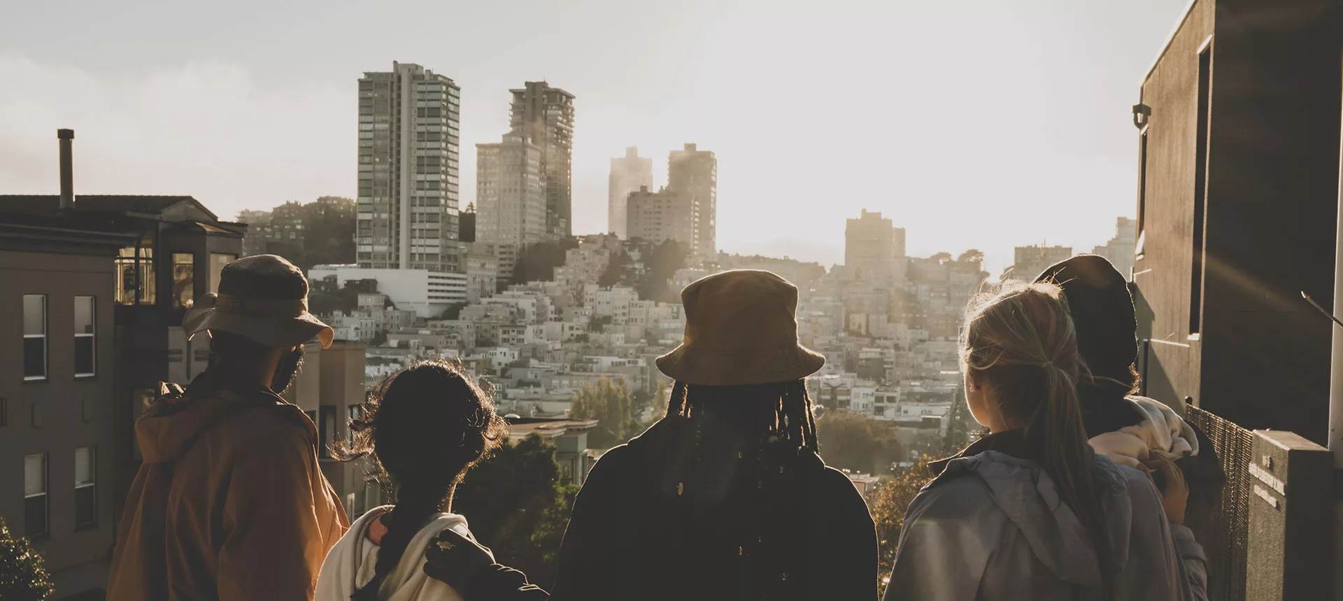 Cinco amigos parados juntos y con chaquetas y sombreros de Timberland, que se muestran desde atrás. Con el sol iluminando sus siluetas mientras miran a una ciudad con edificios blancos.