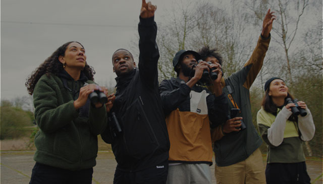 Image of five friends, wearing Timberland, out in a field with trees behind them, all holding binoculars and pointing up.
